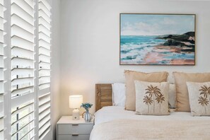 A bedroom with a bed, a nightstand, a lamp, a painting, and a window with blinds. - Sanctuary Splash Getaway (Sanctuary Point)