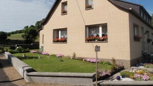 Un edificio de dos pisos con techo inclinado, ventanas rectangulares con macetas de flores y un pequeño jardín con diversas plantas y flores.