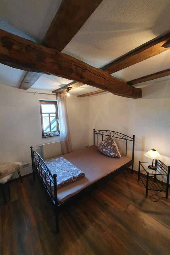 Een kleine kamer met een enkel bed, houten plafond, raam, nachtkastje met lamp en een radiator.