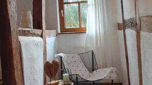 Une pièce chaleureuse avec un plafond orné de poutres en bois, une fenêtre avec des rideaux blancs, une petite chaise en bois, un tapis blanc et moelleux, et un lit avec un cadre en métal.
