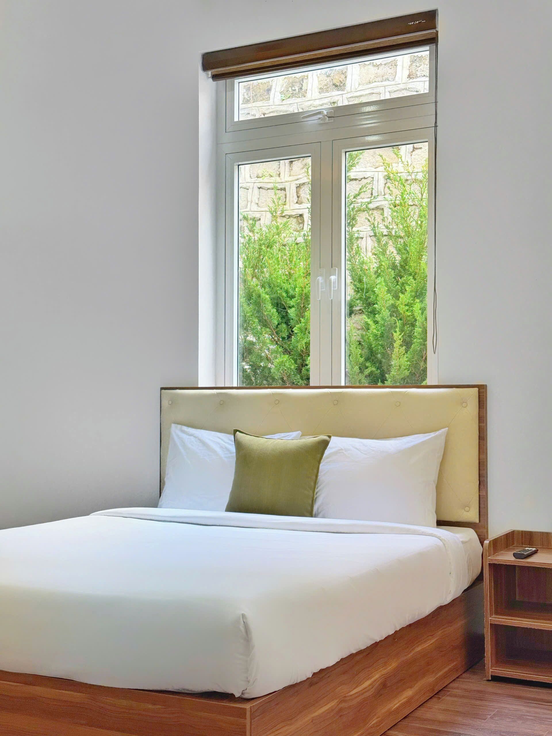 Una cama bien hecha con cabecera de madera, una mesita de noche y una ventana con vistas a la vegetación.