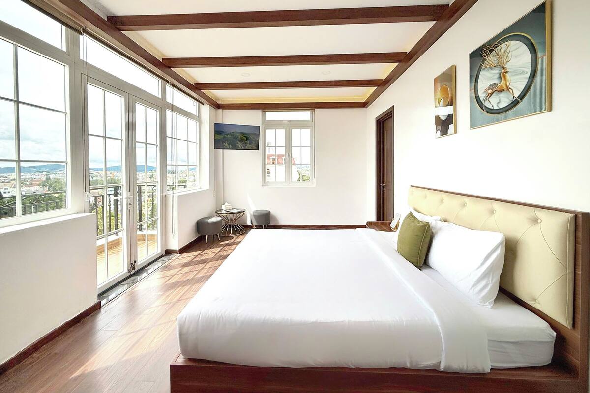 A spacious bedroom with a large bed, wooden beams, a balcony, and a city view.