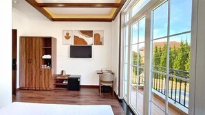 A modern bedroom with a large bed, wooden flooring, a TV, and a balcony with a view of trees and buildings. - KIEN DALAT (Lam Vien - Da Lat)