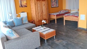 Ein gemütliches Wohnzimmer mit einer Couch, einem Holzschrank, einem kleinen Tisch mit einer Pflanze und einem Bett mit bunter Bettdecke.