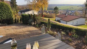 Una terraza de madera con una mesa, una jaula de pájaros y vistas a una zona residencial con casas y árboles.