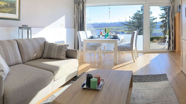 Una sala de estar moderna con un sofá, una mesa baja con velas y un comedor con una mesa y sillas.
