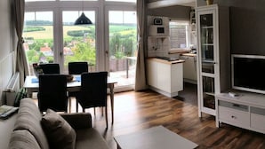 A modern living room with a sofa, dining table, and chairs. There is a kitchen area with a TV and cabinets. Large windows offer a view of the outdoors. - Belle Vue by Interhome (Husen)