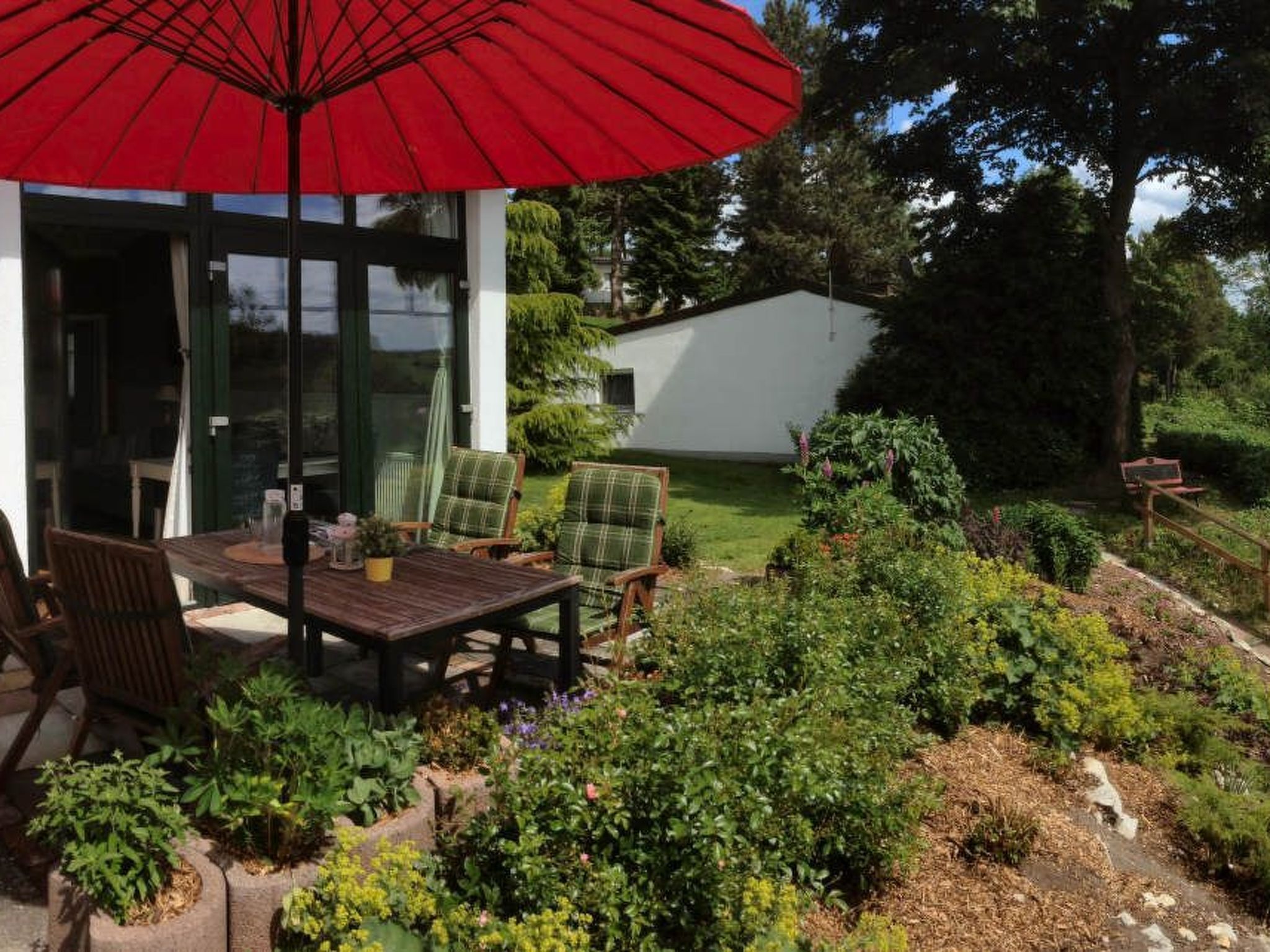 A patio with a table and chairs, a red umbrella, and a garden with various plants.
