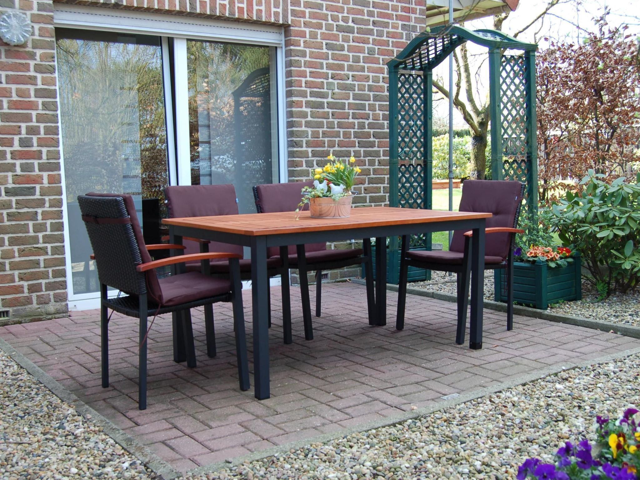 An outdoor patio with a wooden table and four chairs, a brick wall, and a green lattice structure.