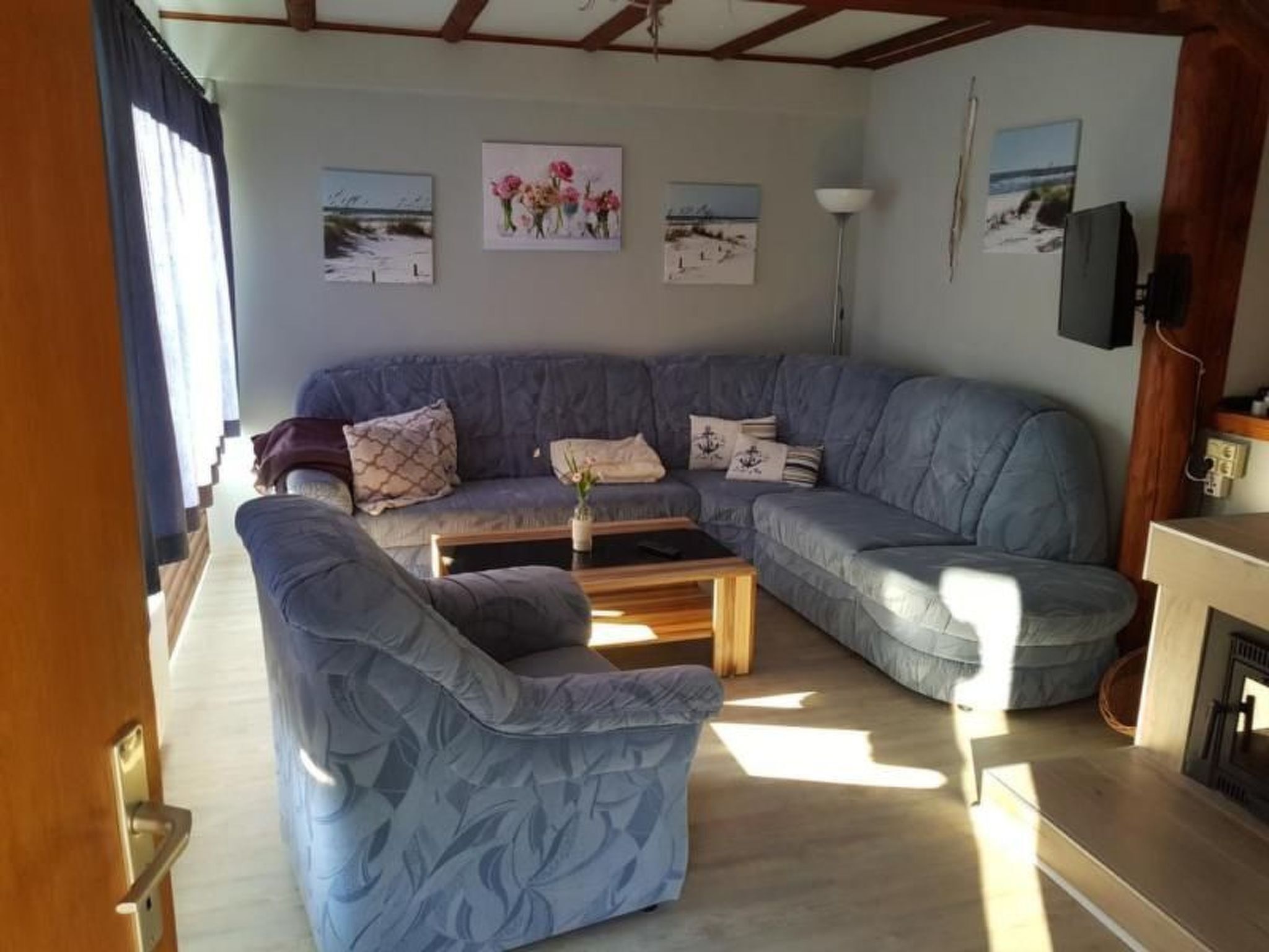 Uma sala de estar com um amplo sofá azul em formato de L, uma mesa de centro de madeira e uma lareira. Há três quadros pendurados na parede, retratando paisagens.