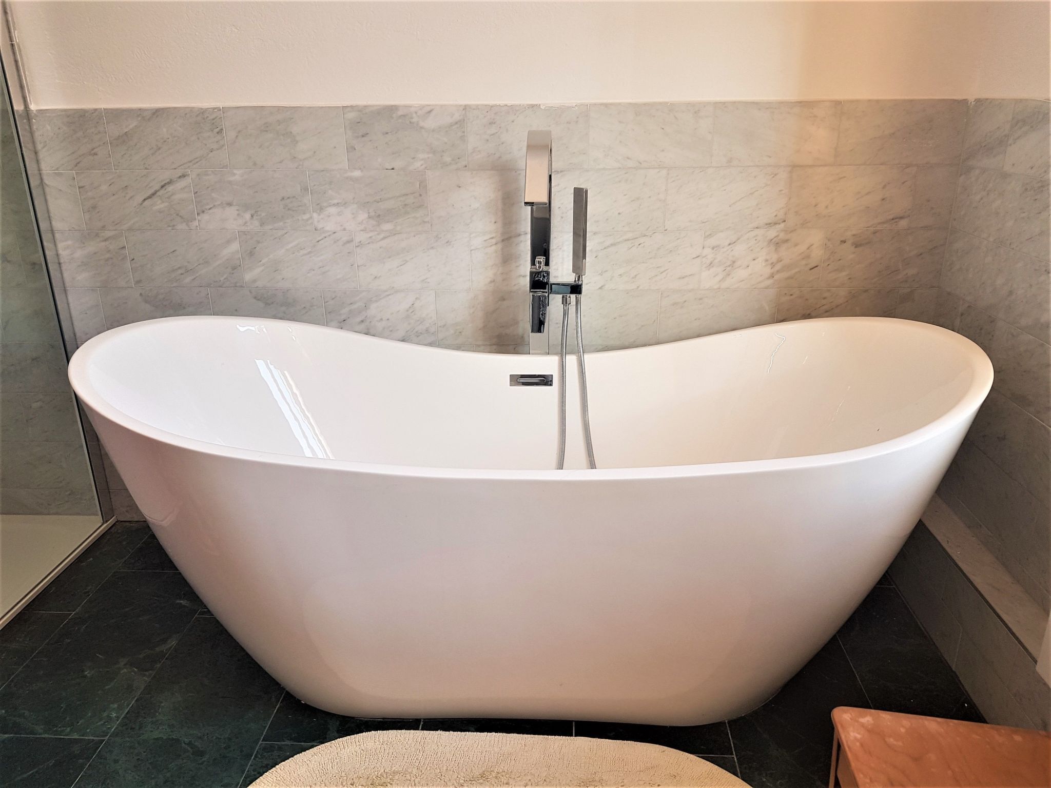 A freestanding bathtub in a bathroom with a tiled floor and a marble wall.