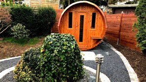 Une structure en forme de tonneau, faite de bois, dotée d’une porte et entourée d’un chemin de gravier et d’une haie.