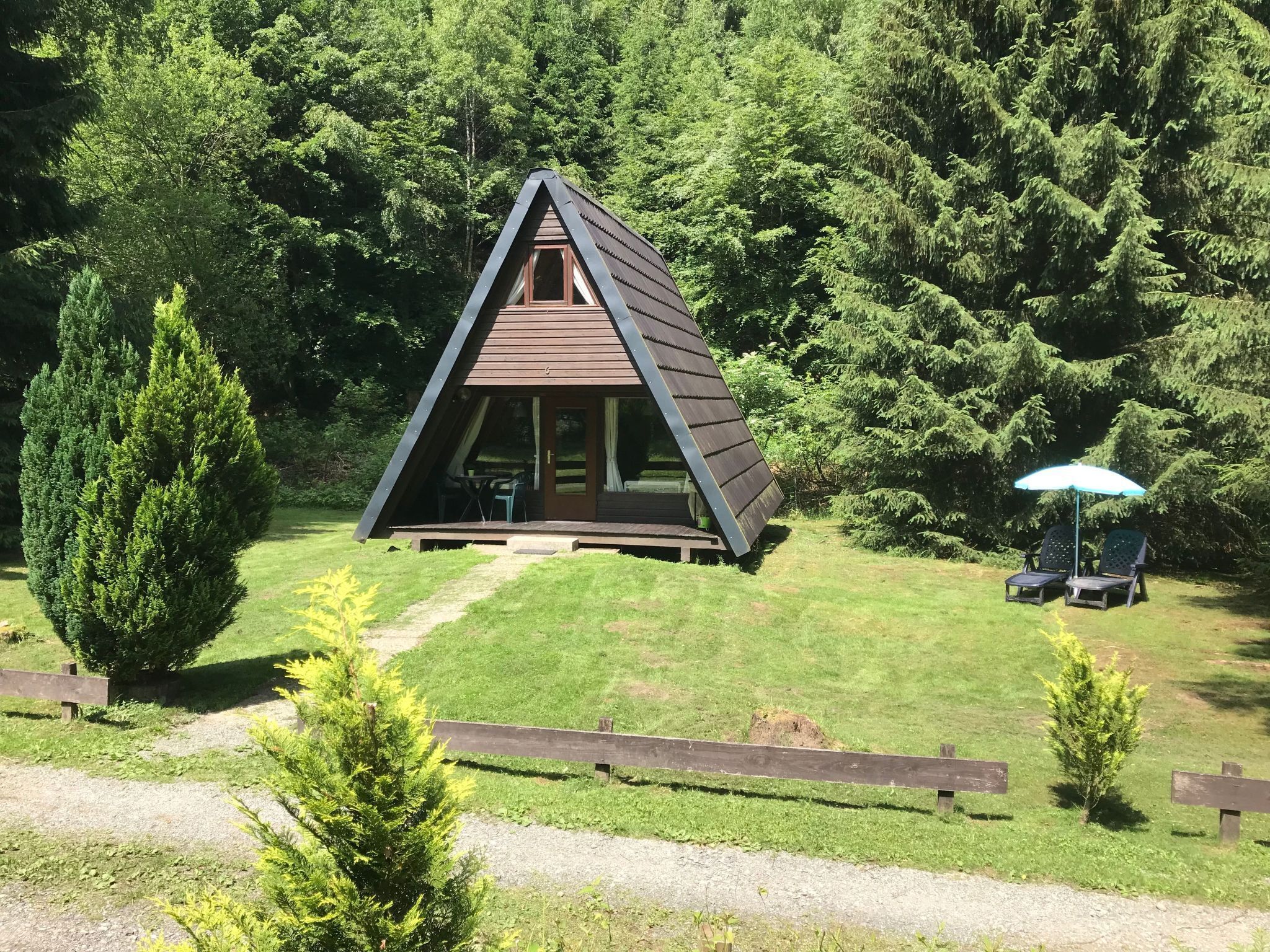 Una pequeña cabaña de madera con forma triangular, puerta y ventanas de vidrio, rodeada de césped y árboles.