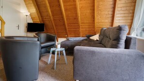 Una habitación estilo loft acogedora, con techo de madera, televisor de pantalla plana en la pared, un sofá gris, un sillón negro y una mesita de vidrio.