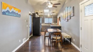 A hallway with a wooden floor, a dining area with a table and chairs, a ceiling fan, and a kitchen area with a refrigerator and window blinds. - Peaceful Bend Escape w/ Luxe Touches, Fenced Yard & Close to Mt. Bachelor & Old Mill, Food and Brews (Bend)
