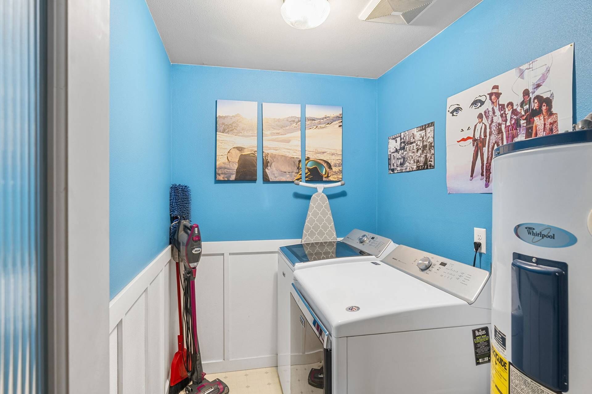 A laundry room with a washing machine, dryer, and iron. There are three framed pictures on the wall and a Whirlpool refrigerator.