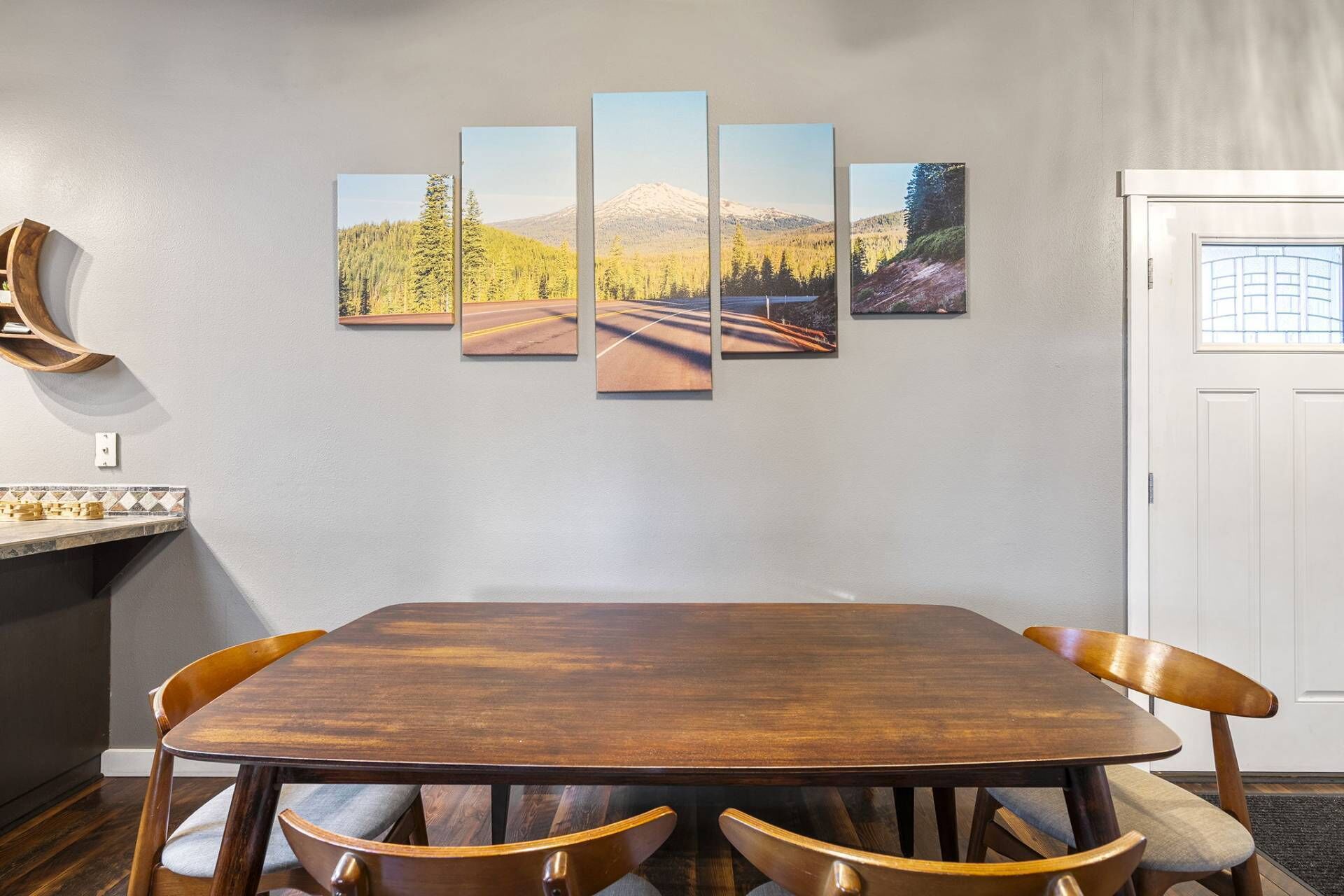 A dining area with a wooden table and chairs, a wall-mounted art piece depicting a mountain landscape, and a white door.