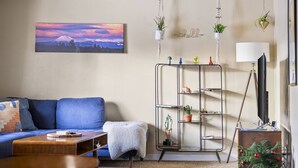 A living room with a blue sofa, a coffee table, a bookshelf with plants, a painting of a mountain, and a television. - Peaceful Bend Escape w/ Luxe Touches, Fenced Yard & Close to Mt. Bachelor & Old Mill, Food and Brews (Bend)
