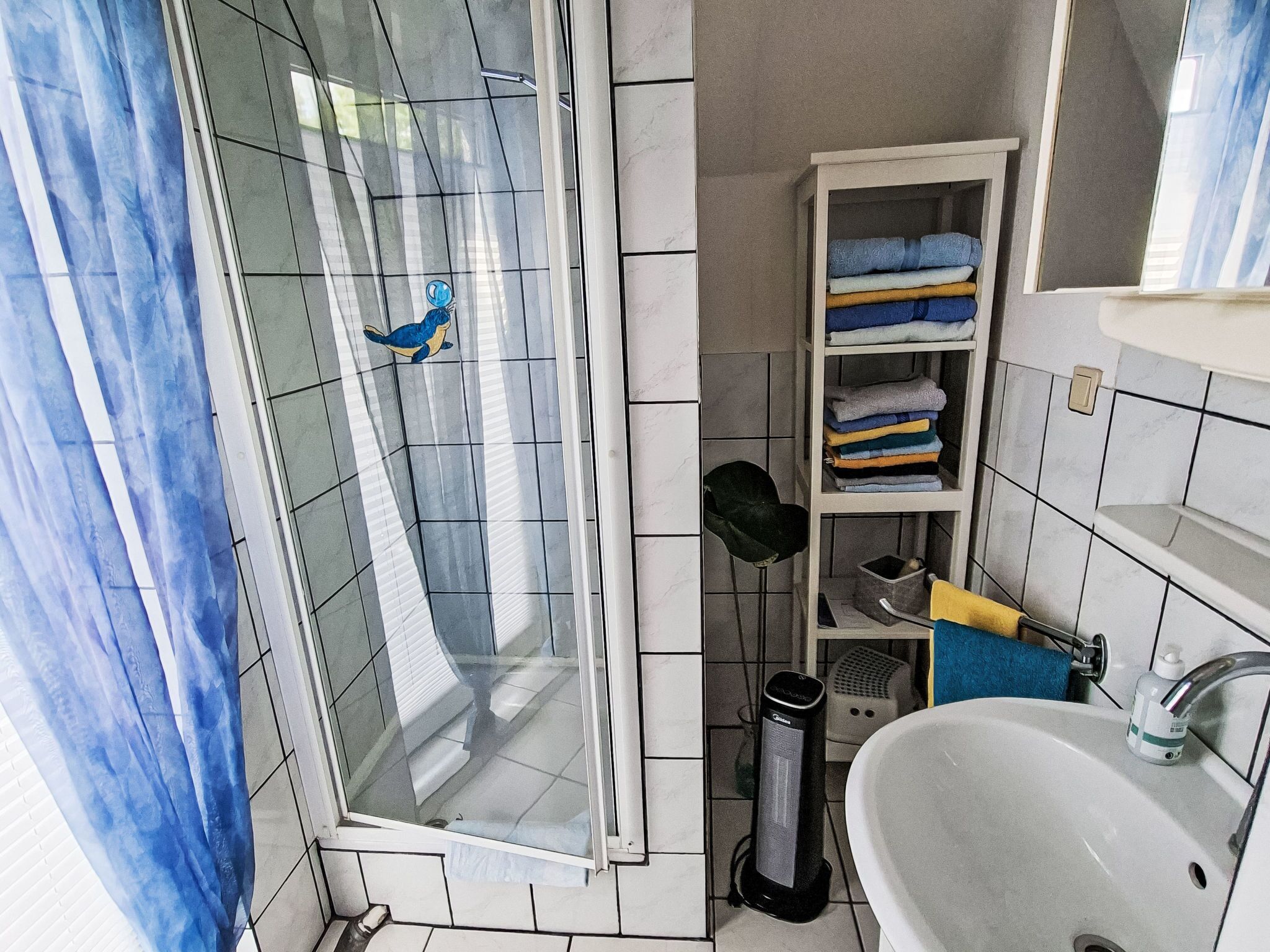A bathroom with a glass shower enclosure, a white sink, a towel rack, and a shelf with folded towels.