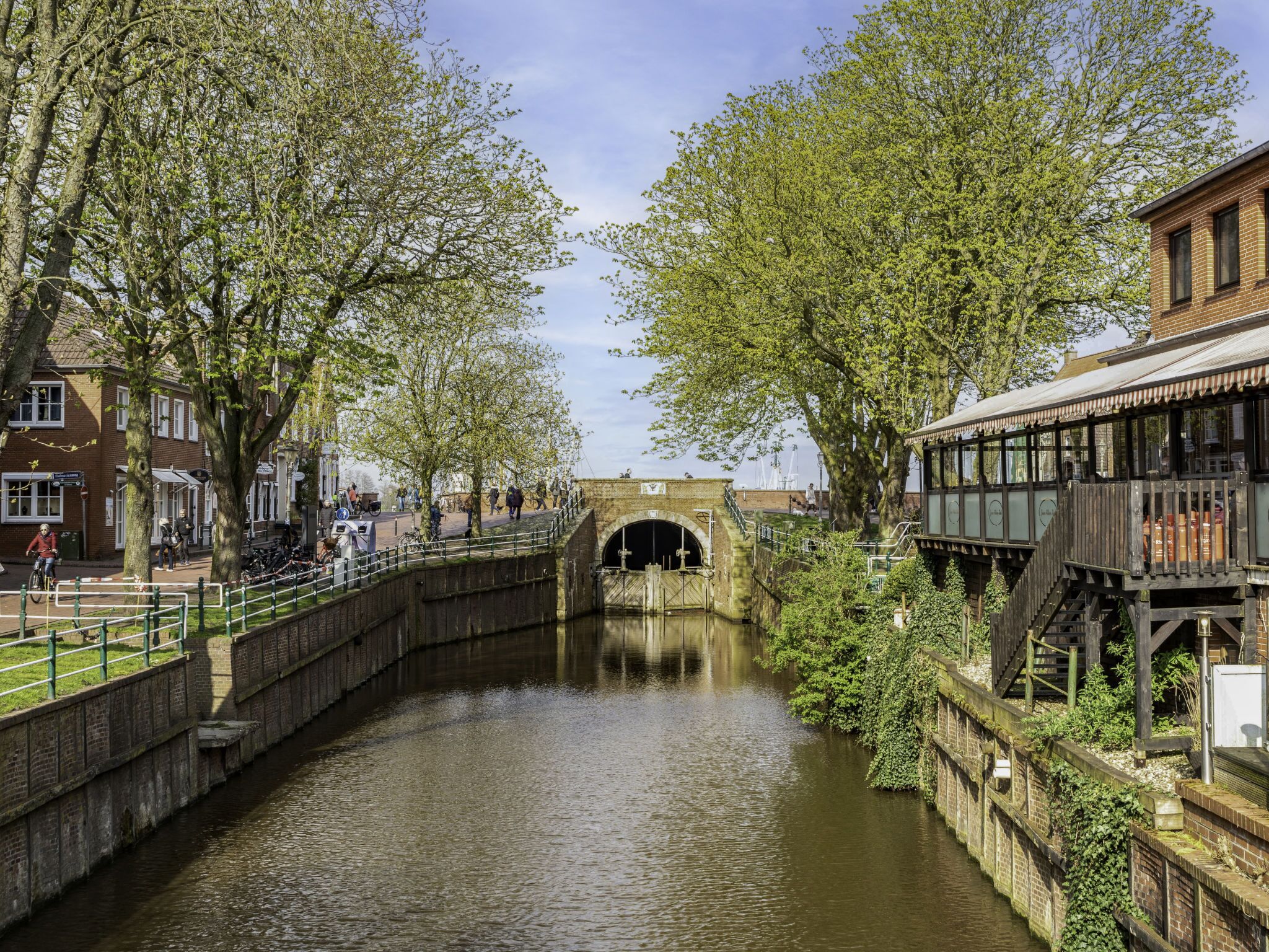 Un canal con un puente de ladrillo, árboles y un edificio con una terraza de madera.