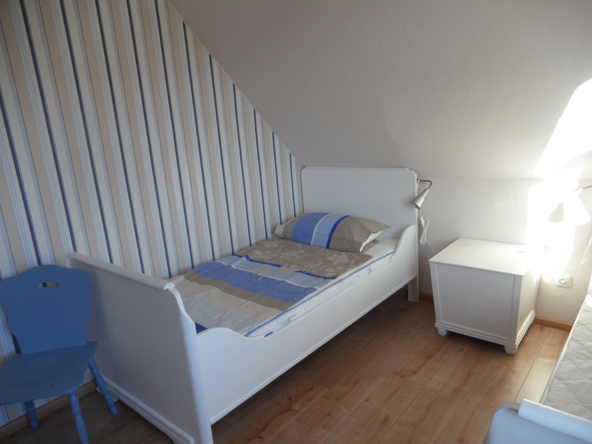 A small, single-bed room with a striped wall, a bedside table, a lamp, and a blue chair.