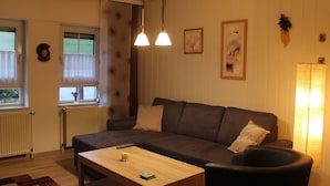 Ein gemütliches Wohnzimmer mit einem dunkelbraunen Sofa, einem Holztisch und einem schwarzen Sessel. Zwei Pendelleuchten hängen von der Decke, ein Heizkörper ist an der Wand angebracht und es gibt ein Fenster mit transparenten Vorhängen.