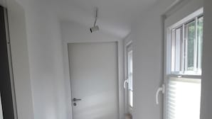 Een gang met een houten vloer, witte muren, een deur aan het einde en een raam met zonwering aan de rechterkant.