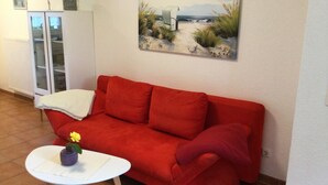 Un sofá rojo con un cojín blanco, una mesita pequeña con un florero, y un cuadro de una playa en la pared.