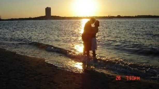 Una pareja abrazándose al atardecer junto al agua, con un edificio a lo lejos.