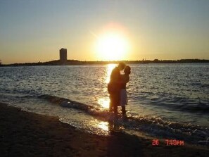 Ein Paar umarmt sich bei Sonnenuntergang am Wasser, im Hintergrund ein Gebäude.