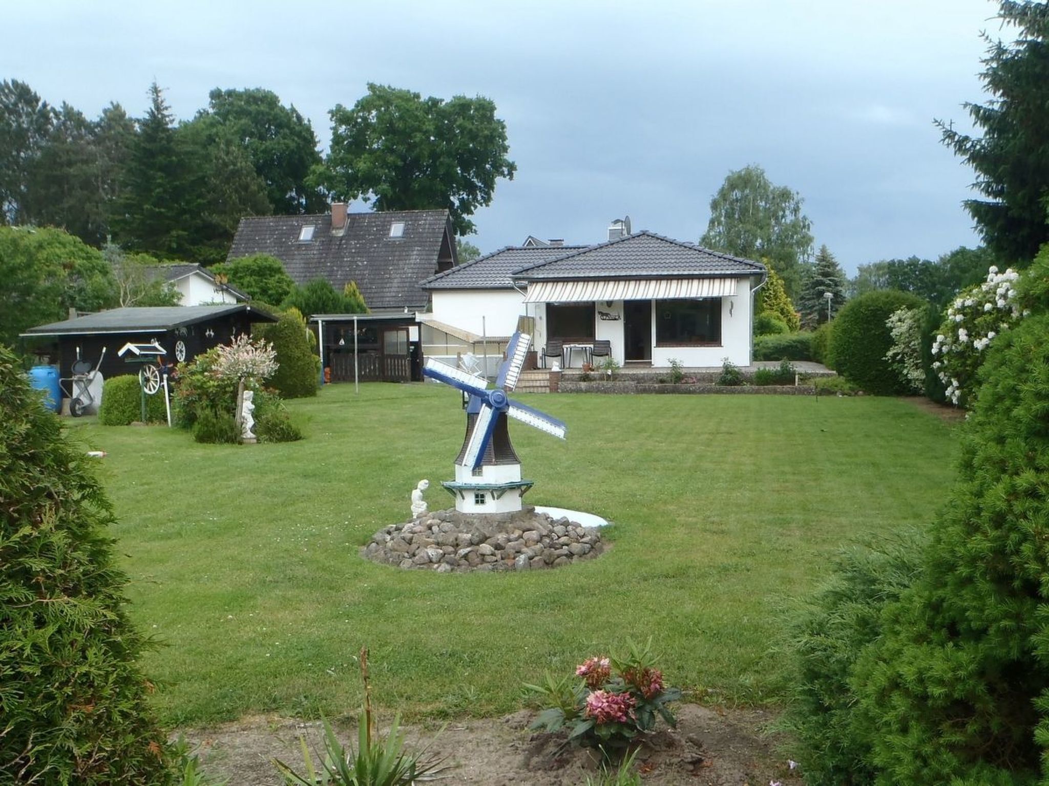 Une maison avec un moulin à vent dans la cour, entourée de verdure et d’un petit jardin.