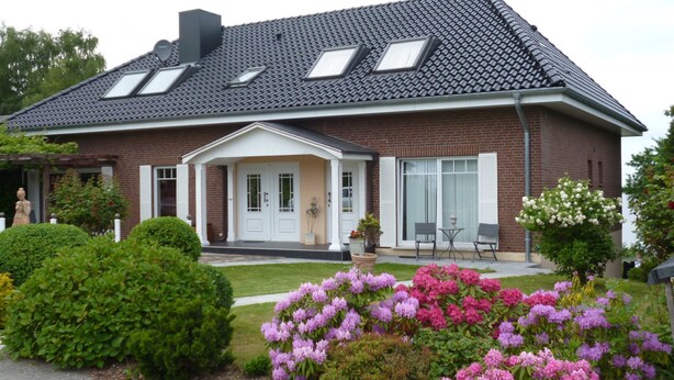 Une maison à deux étages, construite en briques, avec un toit en pente, des lucarnes, un porche à l’avant et un jardin agrémenté de fleurs roses et violettes.