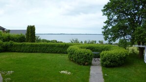 Un jardin bien entretenu, avec un chemin pavé menant à une table et des chaises, entouré de haies soigneusement taillées et offrant une vue sur un lac.