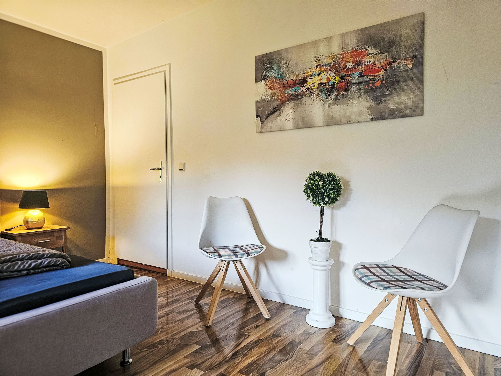 Een slaapkamer met een bed, nachtkastje, lamp, twee stoelen, een potplant en een schilderij aan de muur.