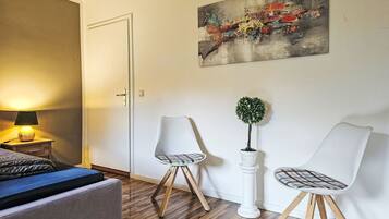 Een slaapkamer met een bed, nachtkastje, lamp, twee stoelen, een potplant en een schilderij aan de muur.