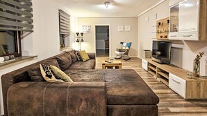 Ein Wohnzimmer mit braunem Sofa, einem Holztisch, einem Fernseher und einem Bücherregal.