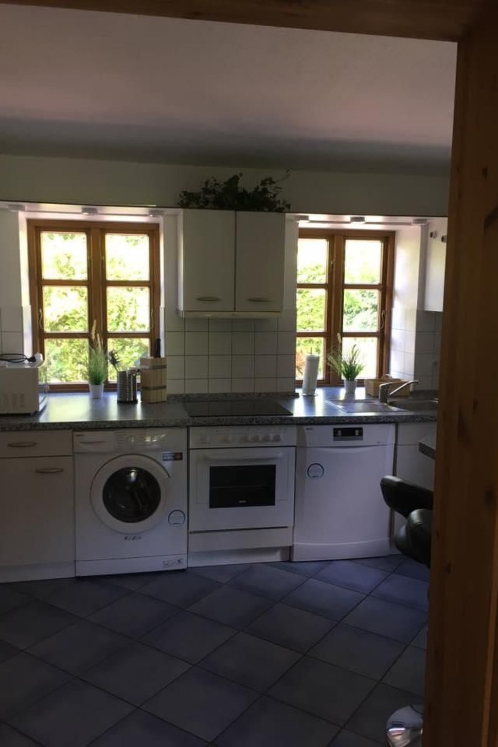 A kitchen with a washing machine, oven, and sink. There are windows with plants on the counter.