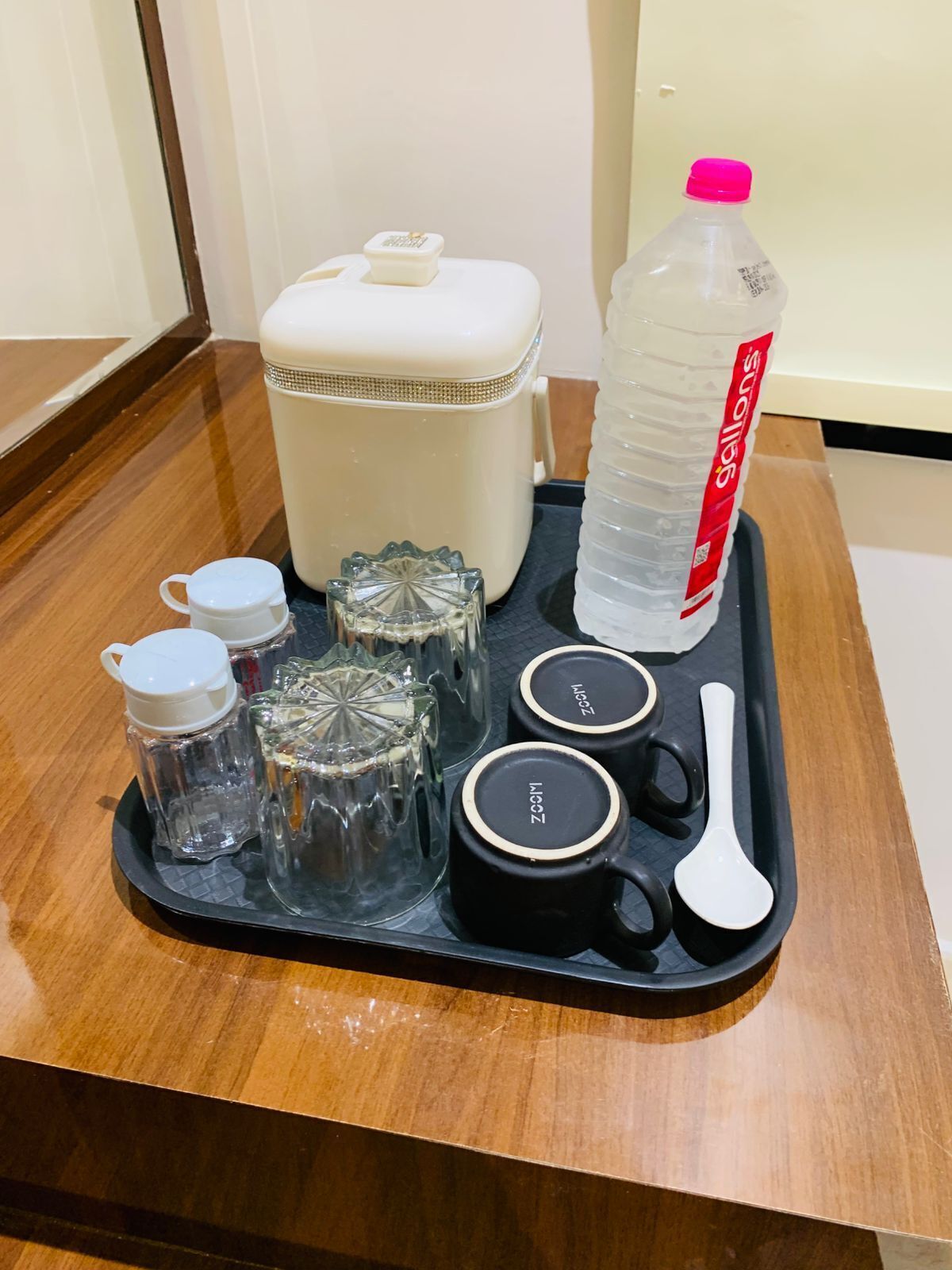Una bandeja con una botella de agua, un recipiente blanco, una taza negra, una cuchara blanca y dos recipientes de vidrio sobre una mesa de madera.