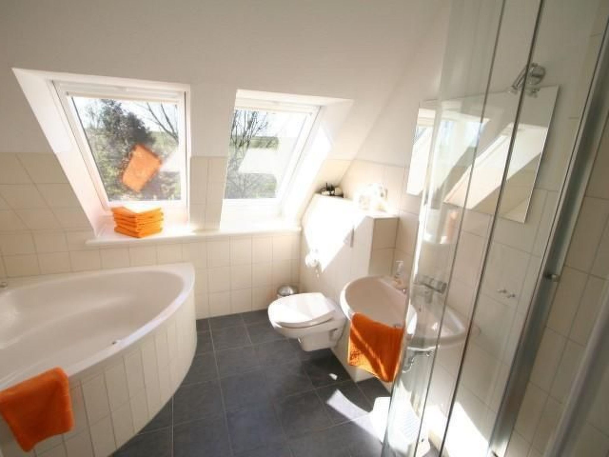 Ein modernes Badezimmer mit Badewanne, Toilette und Duschbereich. Zwei Fenster sorgen für natürliches Licht.
