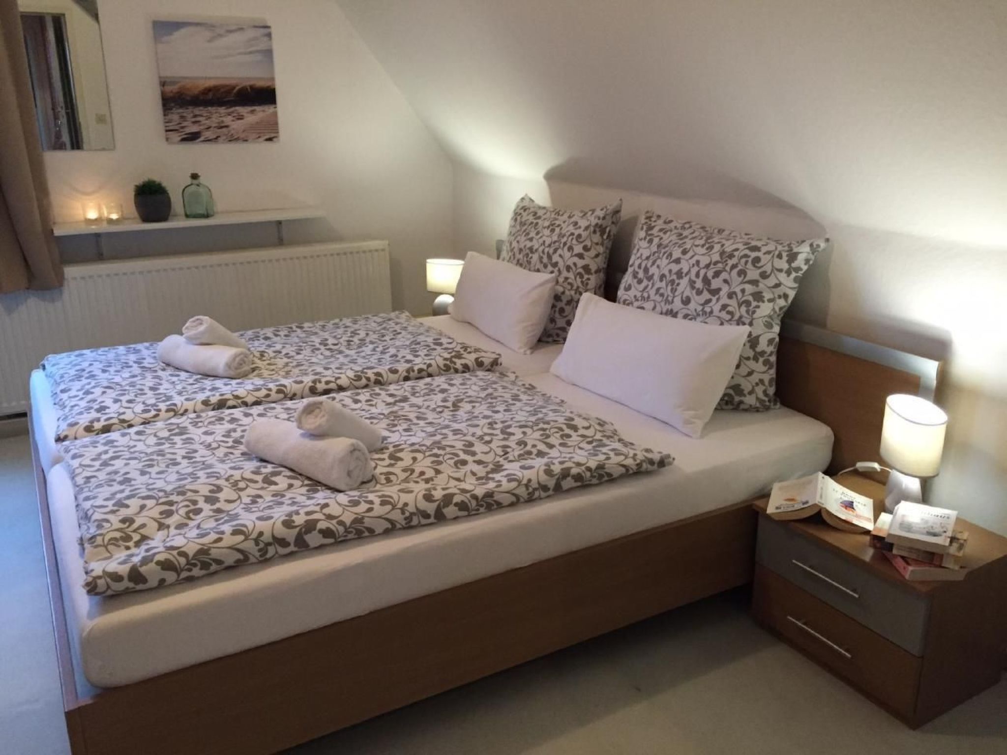 Ein Doppelbett mit gemusterten Betttextilien, zwei Kissen, ein Nachttisch mit Lampe und ein kleines Regal mit Dekoration.