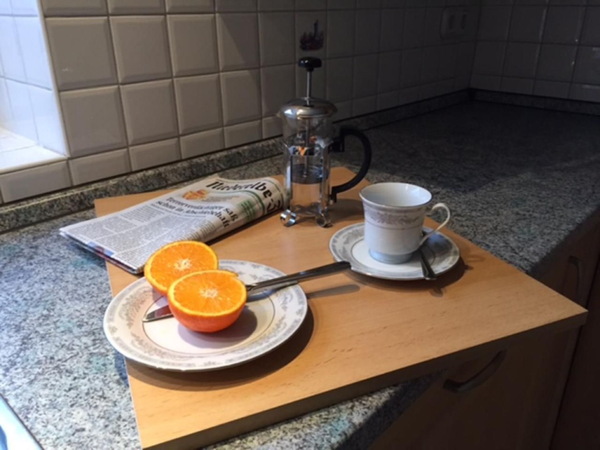 Uma bancada de cozinha com uma cafeteira, uma chávena, um prato com fatias de laranja e um jornal.
