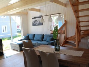 Ein modernes Wohnzimmer mit einem blauen Sofa, einem Esstisch mit Stühlen, einer Treppe und einem großen Fenster mit Blick auf ein Haus.