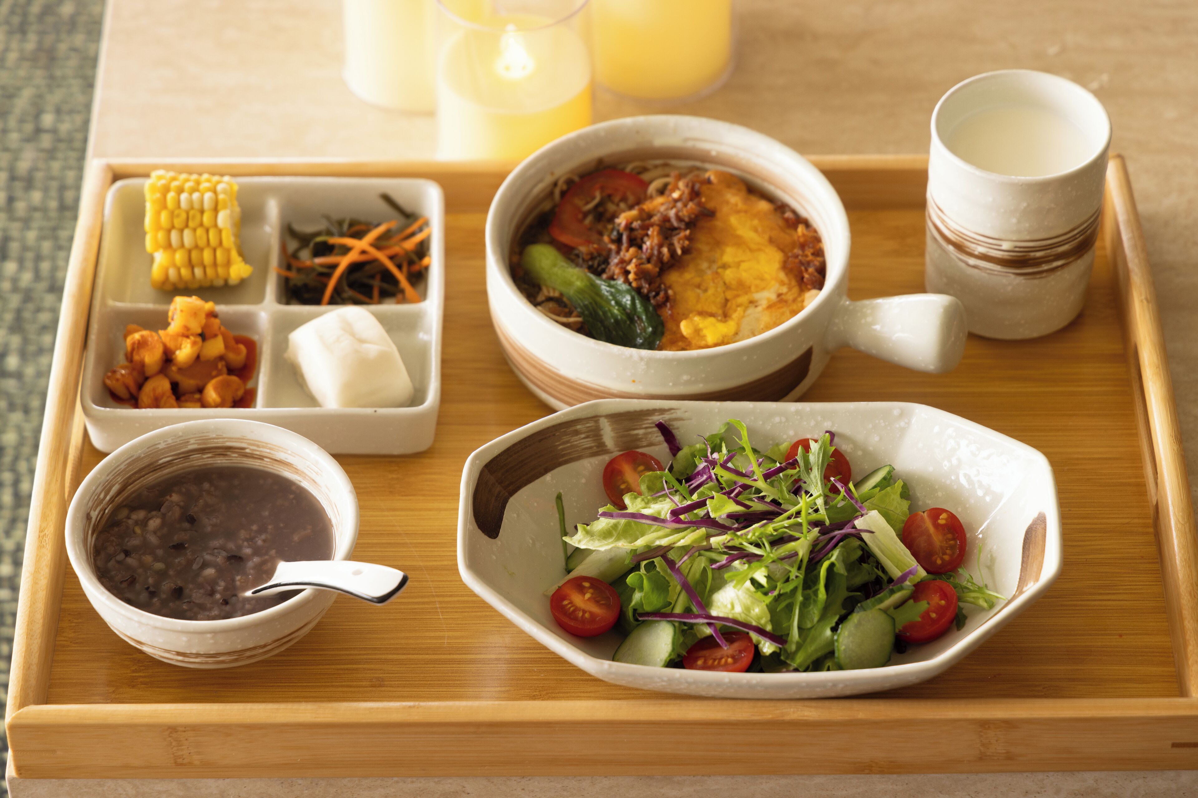 A tray with a bowl of soup, a bowl of salad, a bowl of beans, a corn cob, and a cup of milk.
