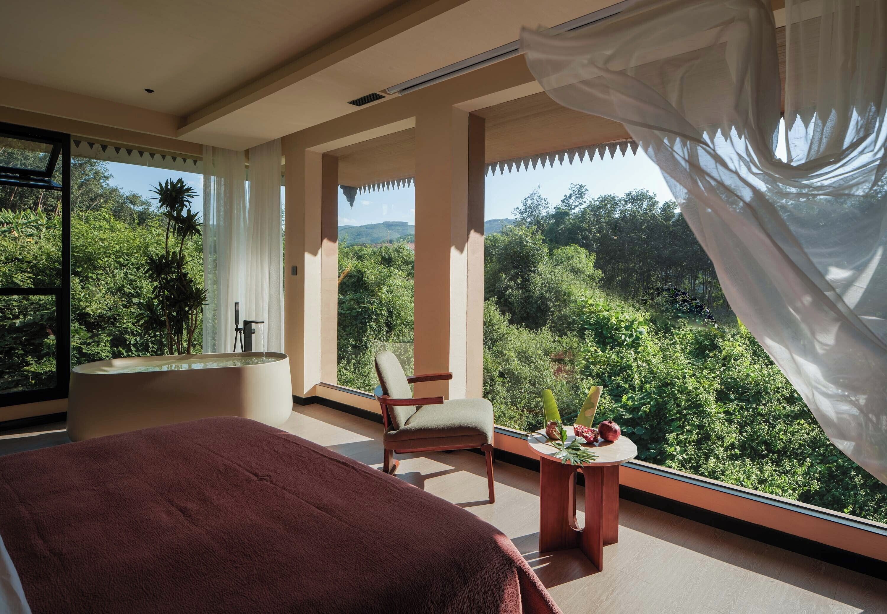 Un dormitorio con una cama grande, una silla, una mesita con flores y vistas a exuberante vegetación a través de amplios ventanales.
