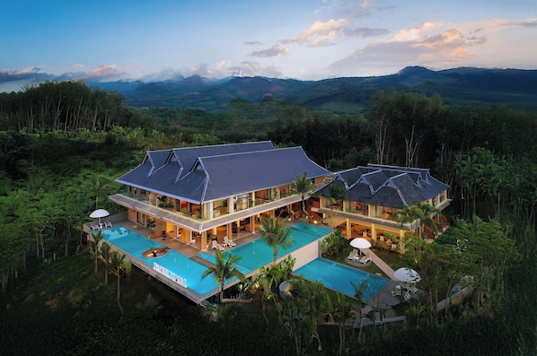A large, multi-story resort with a swimming pool, surrounded by lush greenery and mountains in the background. - Three-Colored Cloud River Resort VILLA (Xishuangbanna Dai)