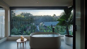 Ein modernes Badezimmer mit einer großen Badewanne, einem kleinen Holzschemel und Kerzen. Es verfügt über ein großes Fenster mit Blick auf einen tropischen Garten und ein traditionelles Haus.