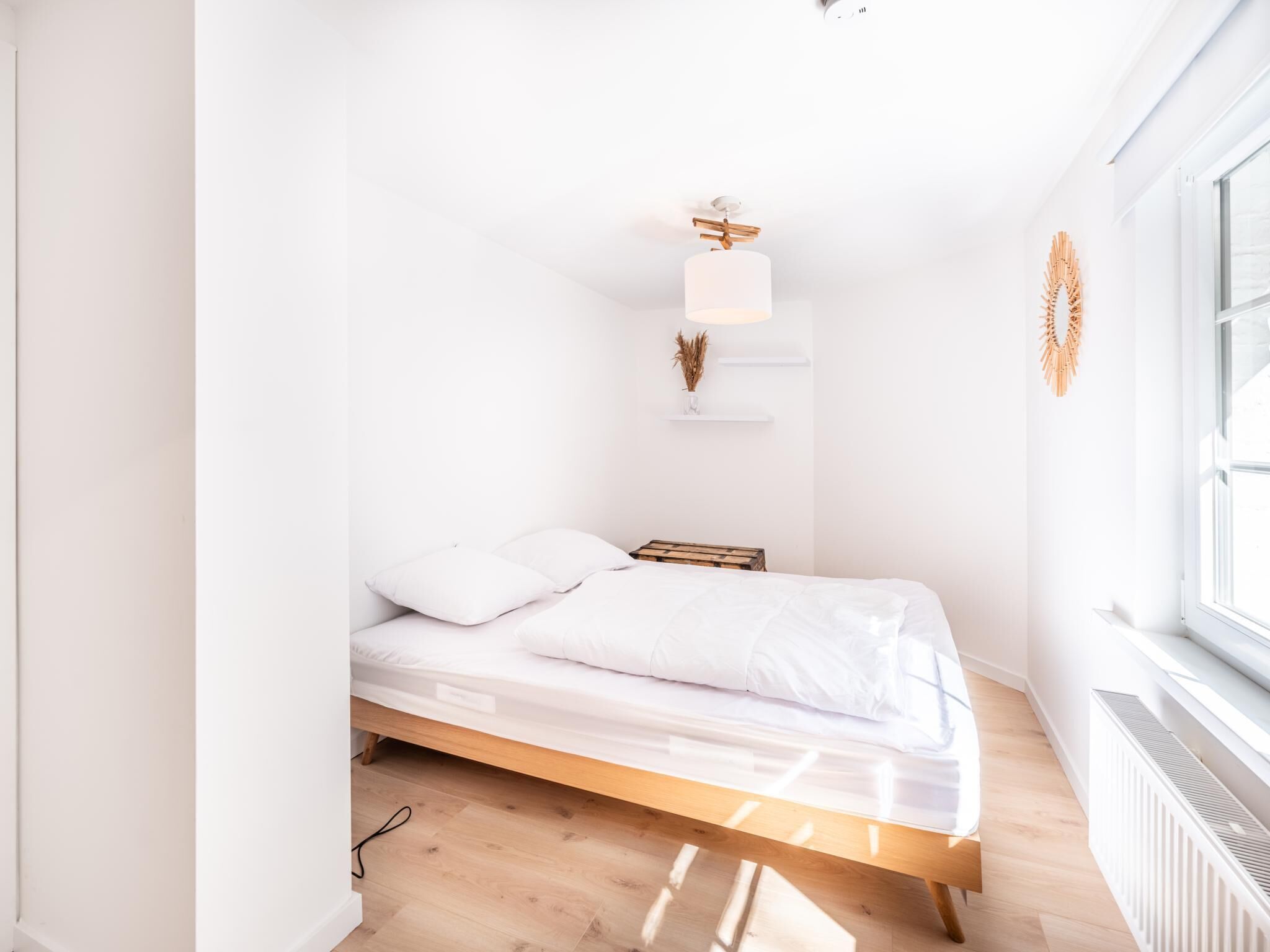 A minimalist bedroom with a wooden bed, white bedding, a bedside table, a wall-mounted shelf with a plant, and a window providing natural light.