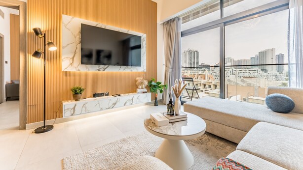 Un soggiorno moderno con una televisione di grandi dimensioni montata su una parete di pannelli in legno, un divano, un tavolino da caffè e vista su uno skyline urbano attraverso ampie finestre.