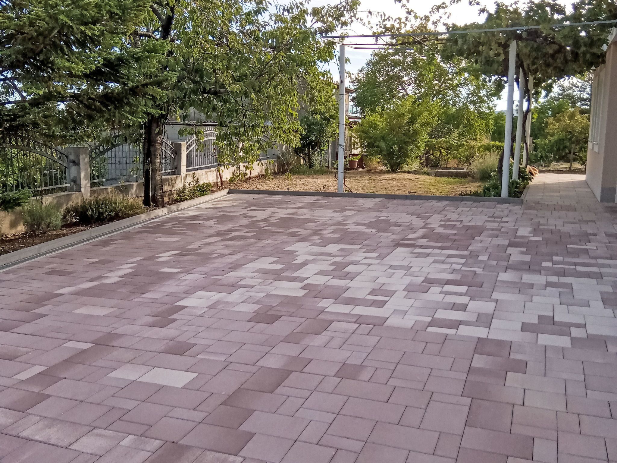 A paved outdoor area with a patterned tile floor, surrounded by greenery and a metal fence.
