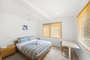 Ein Schlafzimmer mit Bett, Nachttisch, Lampe, Deckenventilator, Kunstwerk, Fenster mit Jalousien und einem Schreibtisch mit Stuhl.